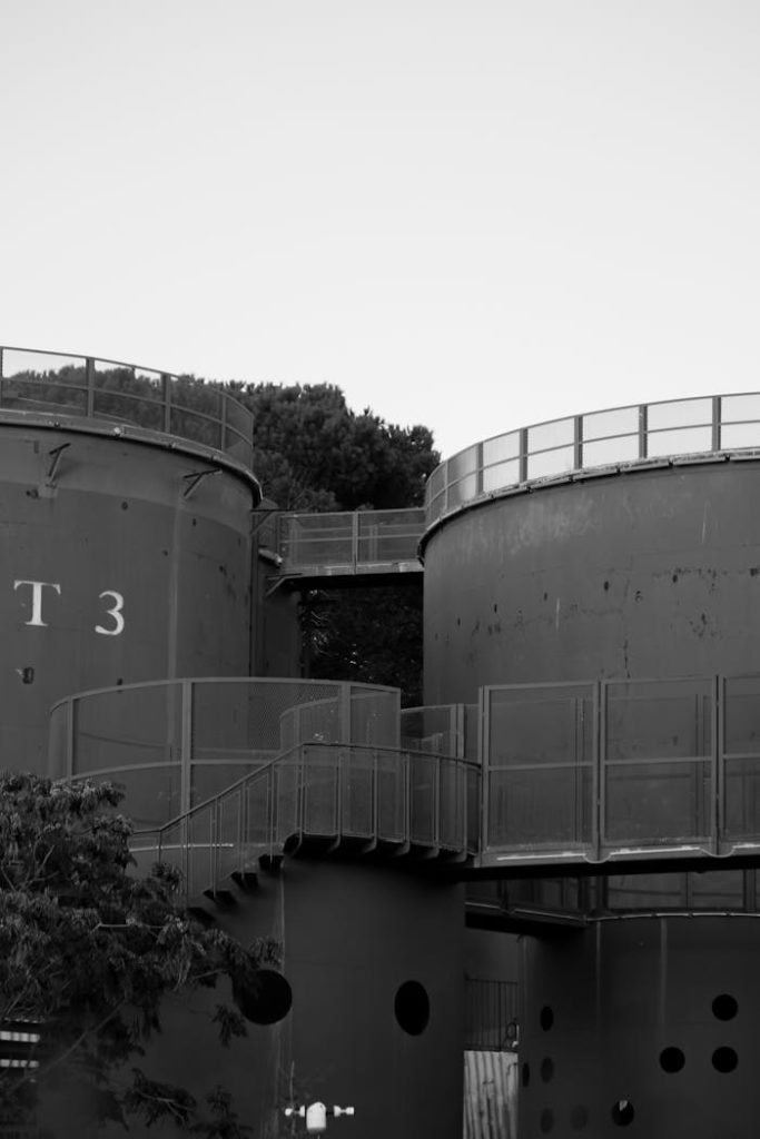 Monochrome photo of large industrial storage tanks with metal railings and trees.