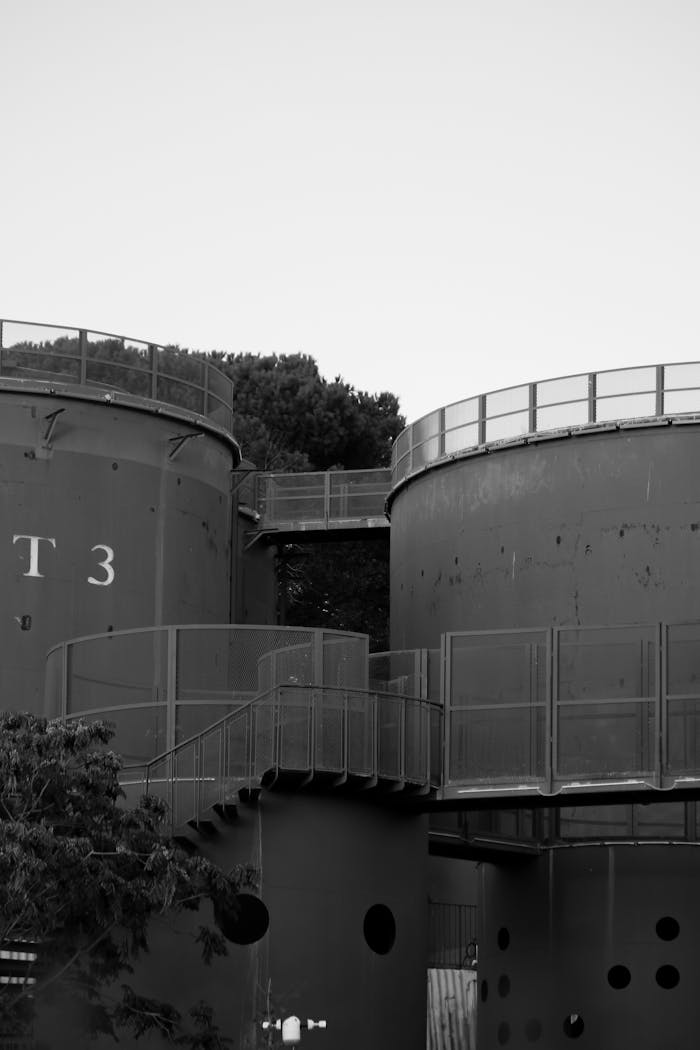 Monochrome photo of large industrial storage tanks with metal railings and trees.